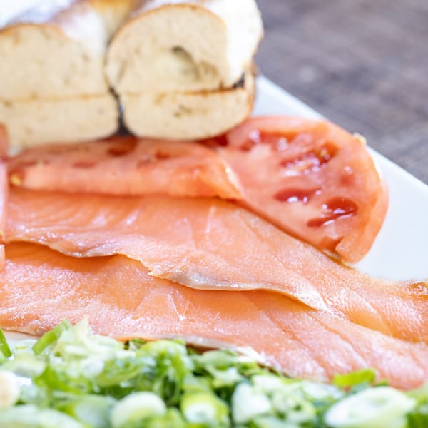Smoked salmon, sliced tomatoes, and green onions on a white plate with a bagel in the background.