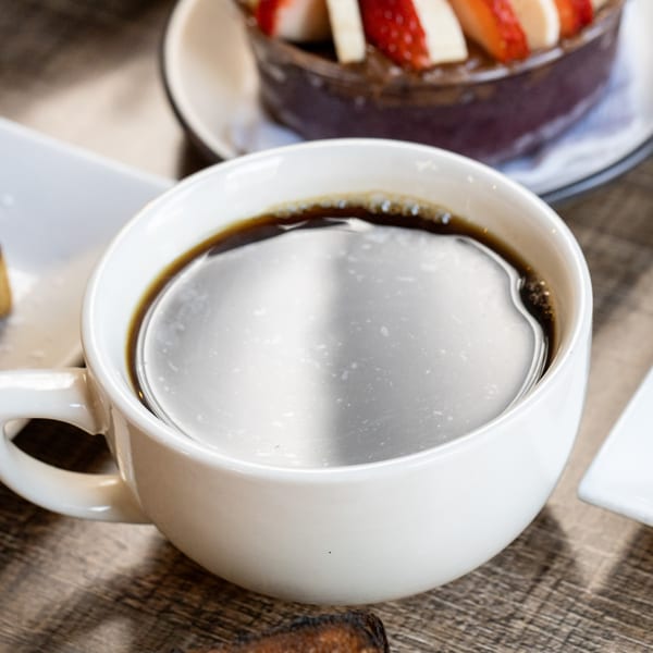 Close-up of a white cup of black coffee on a wooden table, with a fruit bowl in the background.