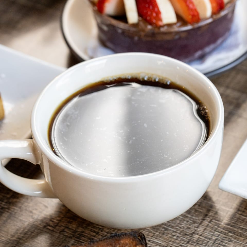 Close-up of a white cup of black coffee on a wooden table, with a fruit bowl in the background.