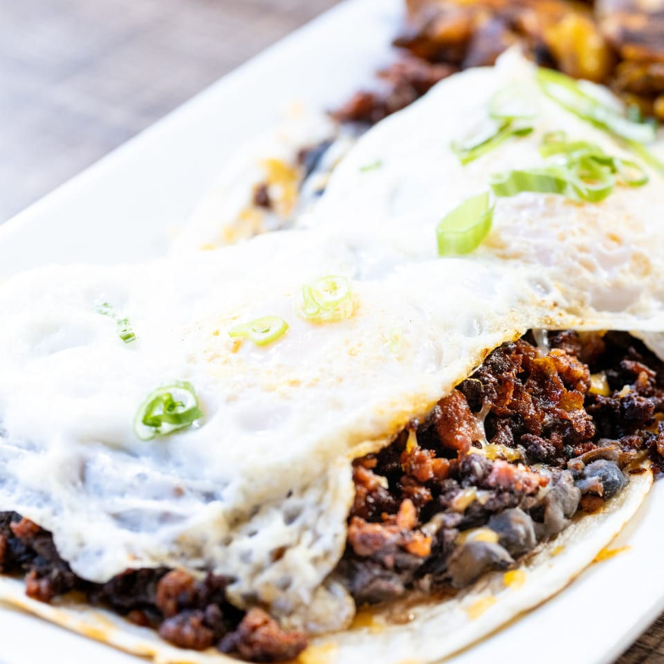 Breakfast tacos with fried eggs, black beans, chorizo, and green onions on a white plate.
